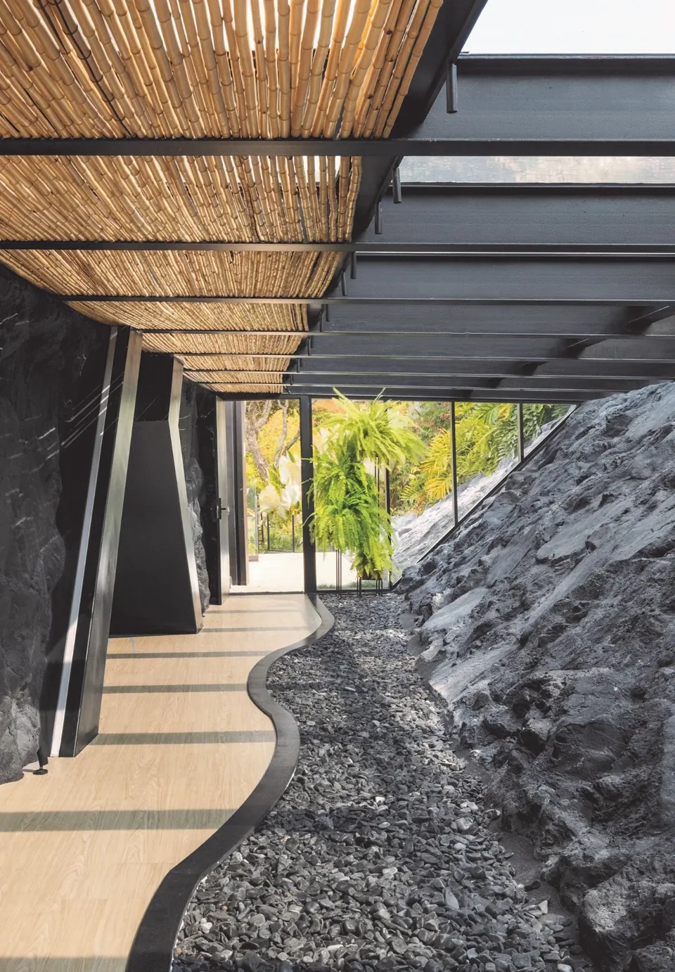 Corredor com parede de rocha em tom carbono e piso de seixos sob a pérgola de madeira